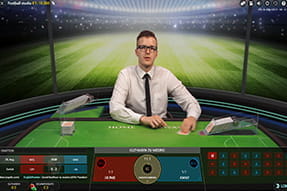 Das Bild zeigt einen Live Dealer in einem fiktiven Fußballstudio, der zwei Stapel Spielkarten in der Hand hält, die jeweils eine Fußballmannschaft simulieren.